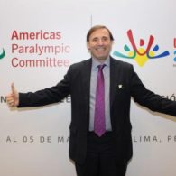 A man holds his arms open in front of a banner for the Americas Paralympic Committee