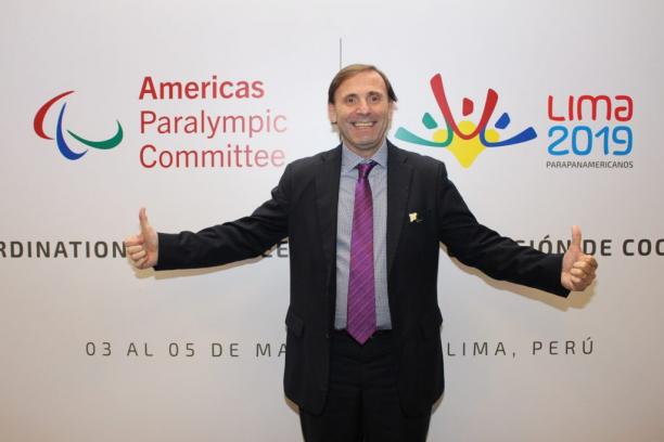 A man holds his arms open in front of a banner for the Americas Paralympic Committee